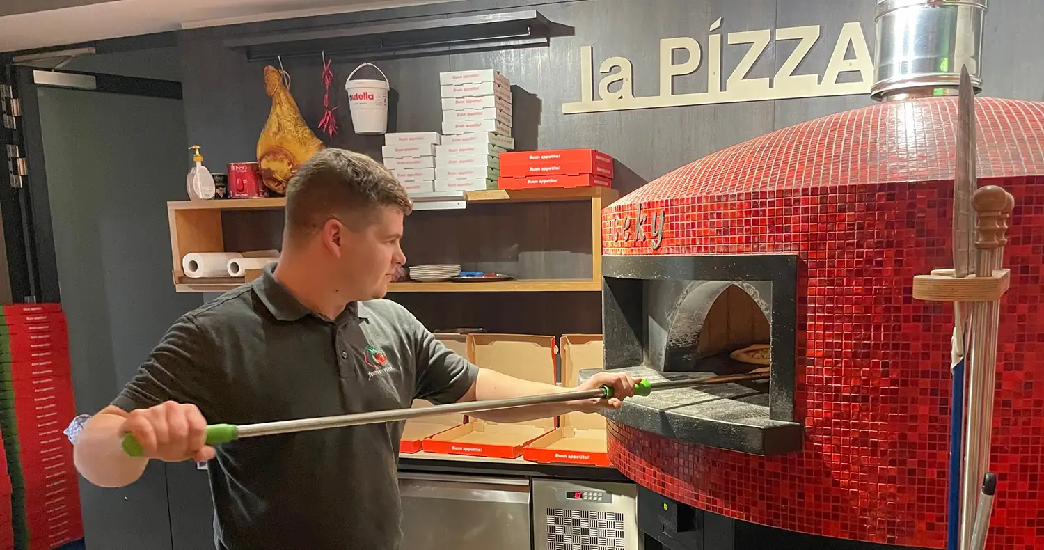 Junger Pizzabäcker mit Ristorante Pomodorino T-Shirt, der gerade eine Pizza aus dem Holzbackofen holt. Im Hintergrund Pizzaschachteln und ein Schriftzug la Pizza